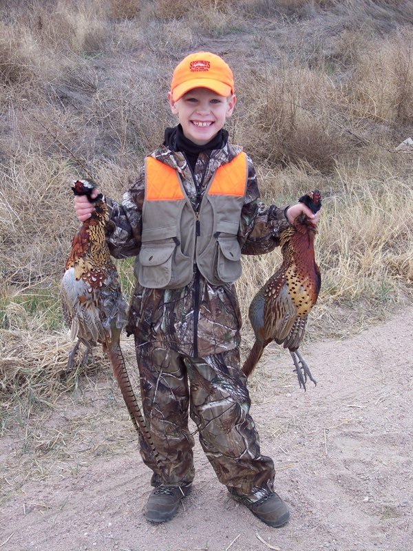 Young hunter in camo and a blaze-orange cap and vest holding two rooster pheasants